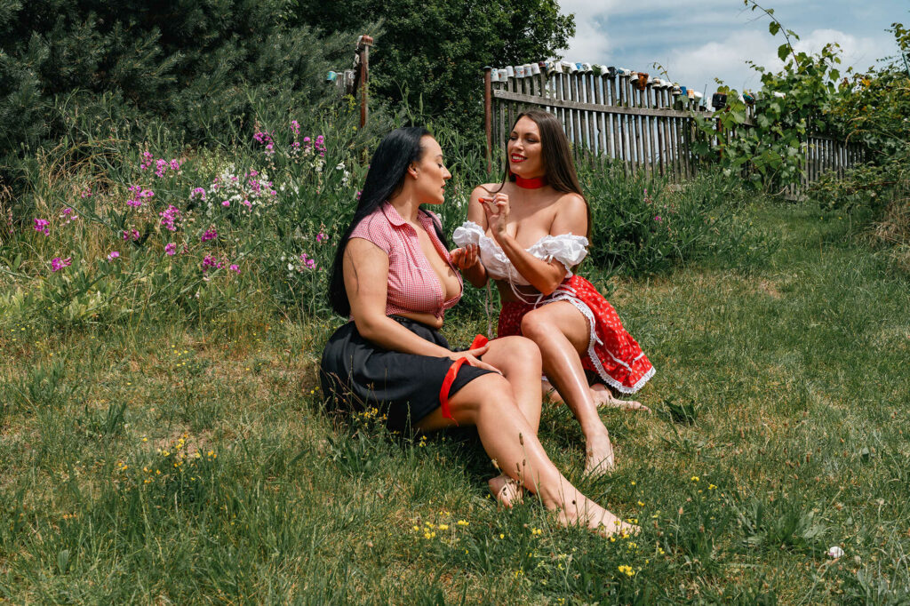 Two young women in folk costumes sitting in the grass, picking and eating fresh red currants together, HotFolk calendar