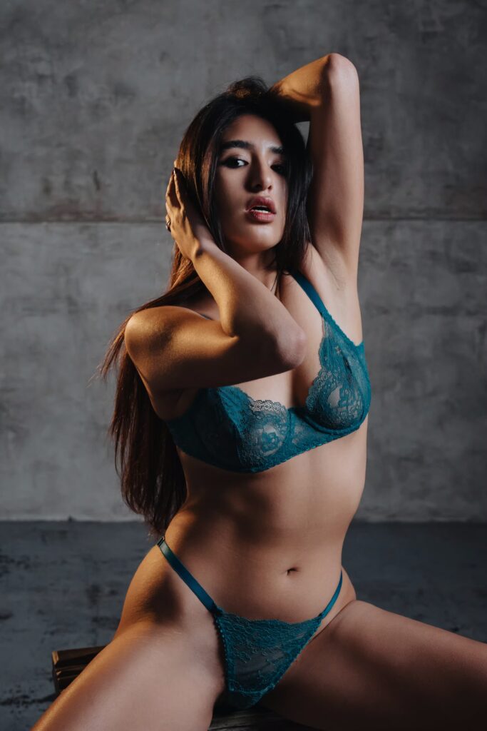 Sofia in blue lace lingerie with her hand in her hair against a grey wall