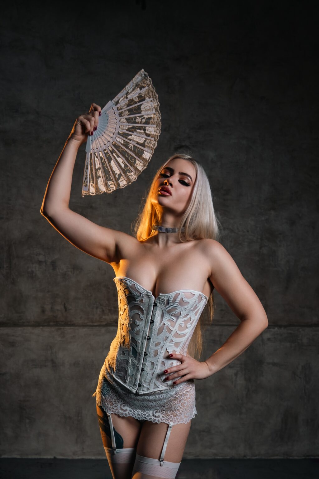 Vermilion in a white corset holding a fan in a dark studio