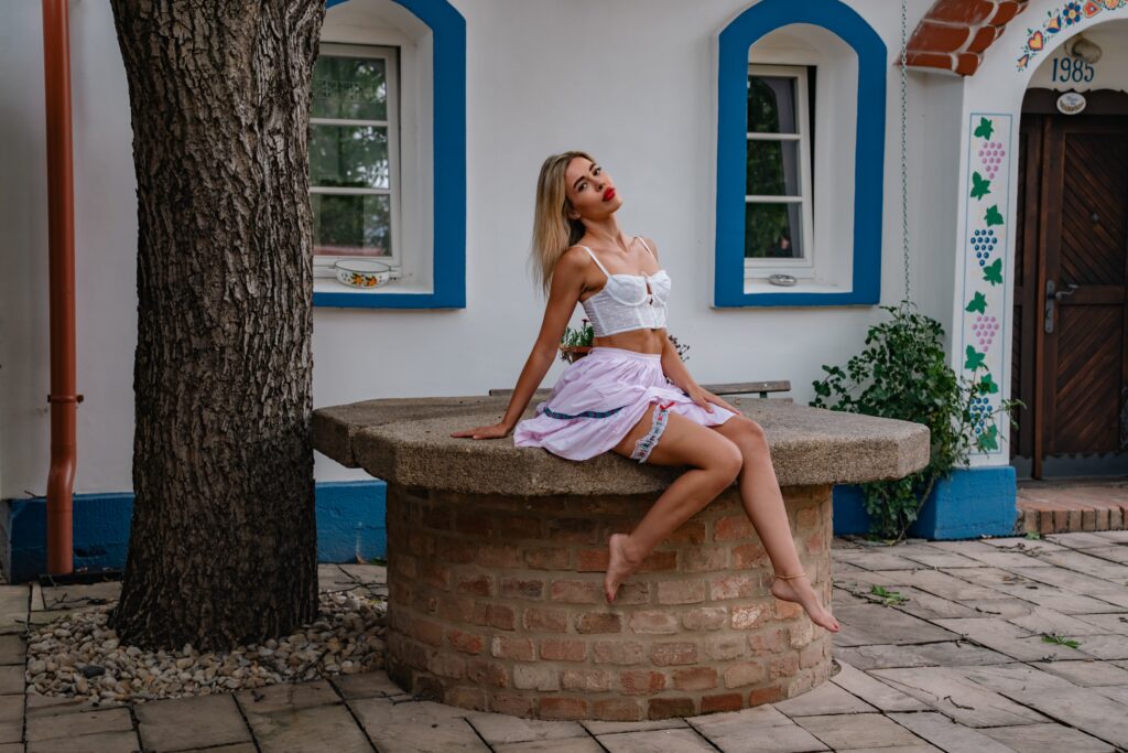 A young woman sitting by a water pump in the morning, posing naturally with her long legs during a HotFolk photoshoot.