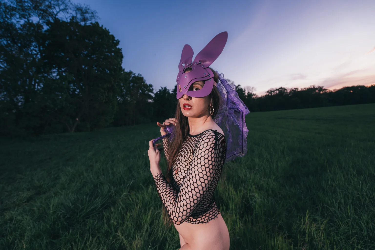 Anna in a purple bunny mask and fishnet top posing in a green meadow