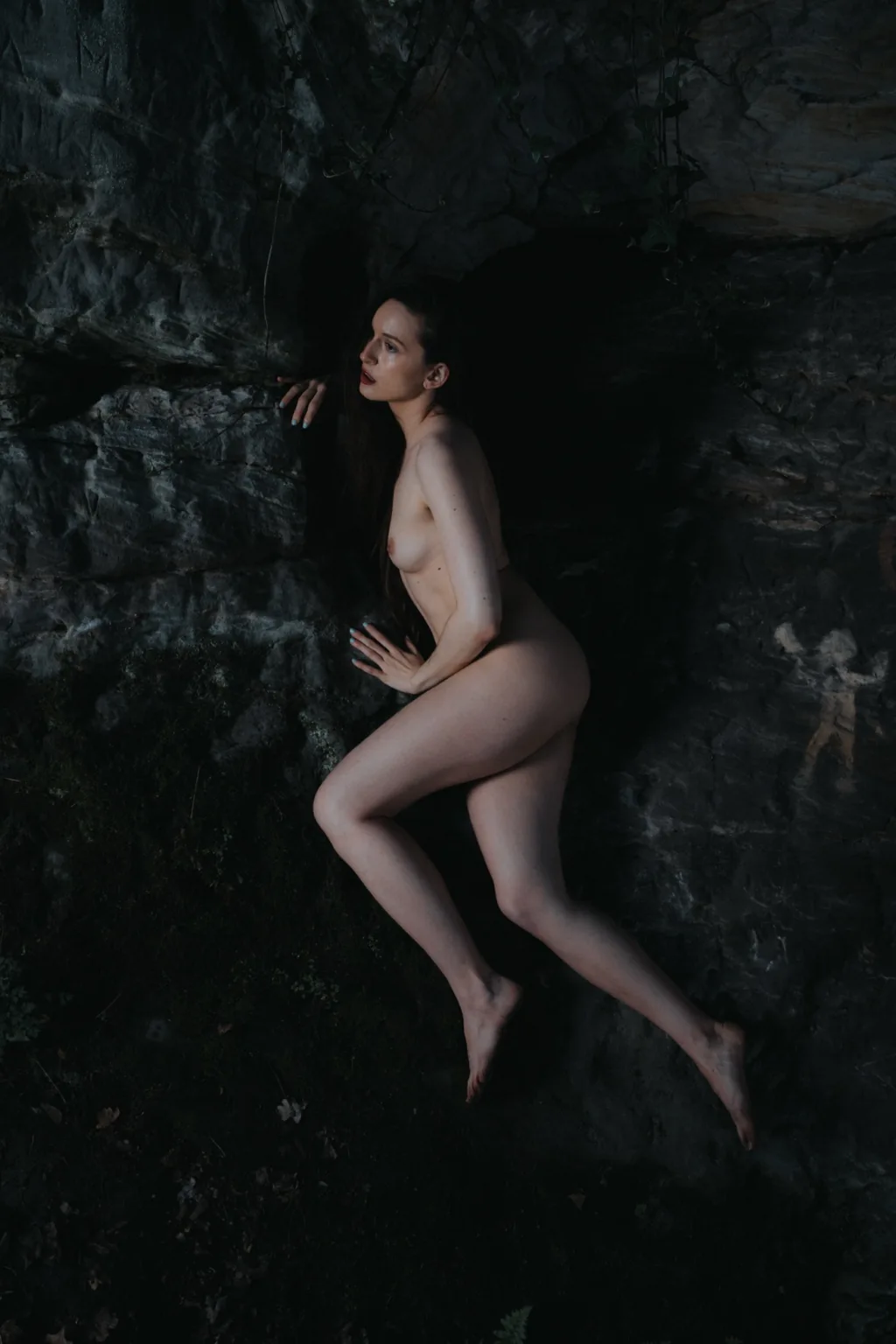 Anna posing nude against a dark rock wall in the forest
