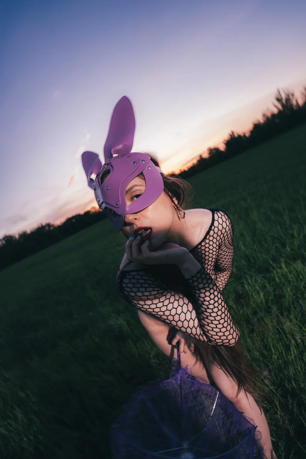 Anna in a purple bunny mask and fishnet top in a meadow at dusk